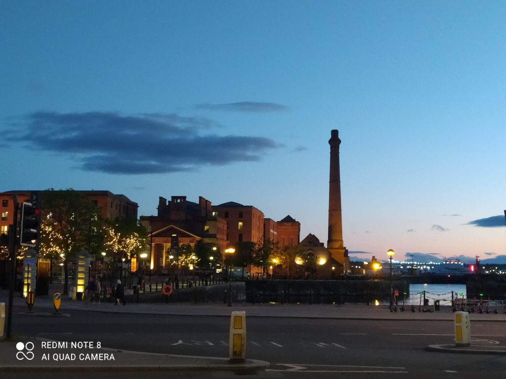 Photograph of the Albert Docks in Liverpool, UK at sunset, showing buildings to the left with the pump house tower with the road at the forefront and the sky deep blue tinged with red and a single cloud in the sky