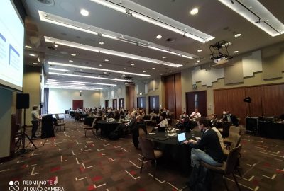 Photograph from the I2I May 2022 Convening showing a large conference room with people sitting at round tables with a speaker presenting at a podium and a presentation on a screen behind the podium.