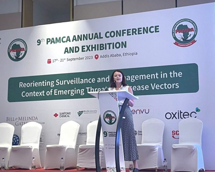 A woman with brown short hair, wearing a white short sleeve shirt and blue trousers standing at a white podium with the PAMCA logo on it on a stage in front of a PAMCA banner and a row of white chairs.