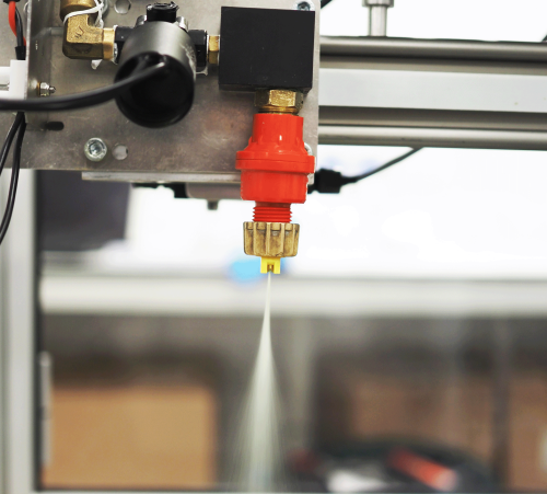 Image of the Micron Horizontal Track Sprayer spray nozzle in action. In the centre of the image is a red and gold nozzle on a silver and black metal track that is horizontal across the image. A fine stream of liquid is spraying downward from the nozzle. Behind the nozzle is a glass window with a silver metal frame.