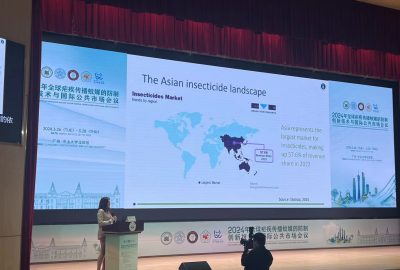 Large screen above a stage where a woman stands at the left of the screen at a podium. The screen displays a global map with the title 'The Asian insecticide landscape'.