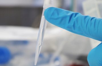 Pipette tip with clear liquid at centre of image, held in place by an index finger in a blue latex glove