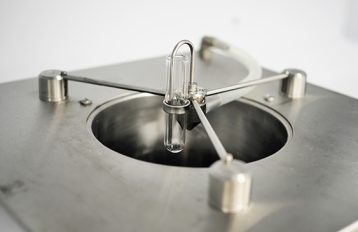 Image of the top of the Potter Tower. Metal, silver platform with a deep, circular hole in the centre. A clear, glass tube is suspended above the hole and held in place by three equidistance silver, metal rods attached to the platform. A thin, u-shaped, inverted metal tube rests within the glass tube and a clear rubber tube is attached to the metal tube and runs to the back of the platform at the back of the photo.